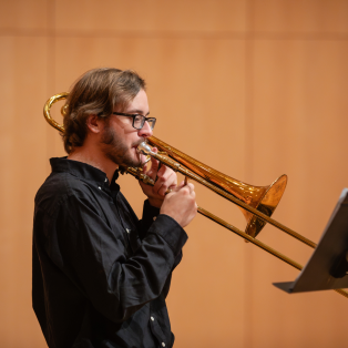 A person with glasses plays a brass trombone while standing in front of a music stand. The background is a soft, tan-colored wall. They wear a black shirt and appear focused on the music.