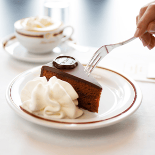 A hand holds a fork poised to cut into a slice of chocolate-covered cake with a whipped cream garnish on a white plate; a cup of coffee sits in the background.