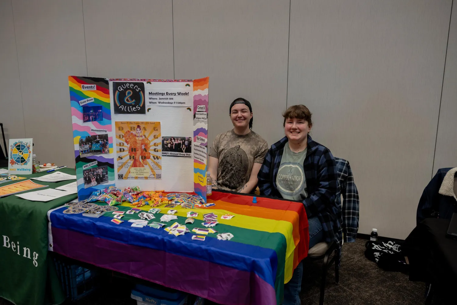 Two people sit at a table covered with a rainbow pride flag, promoting an LGBTQ+ group with flyers, buttons, and candy. A colorful display board stands behind them in a room with neutral-colored walls.