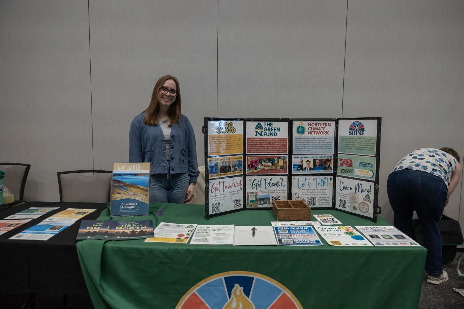 A woman stands behind a display table with posters about The Green Fund, sustainability, and student involvement at an indoor event. Another person is bent over, arranging items on a nearby table.