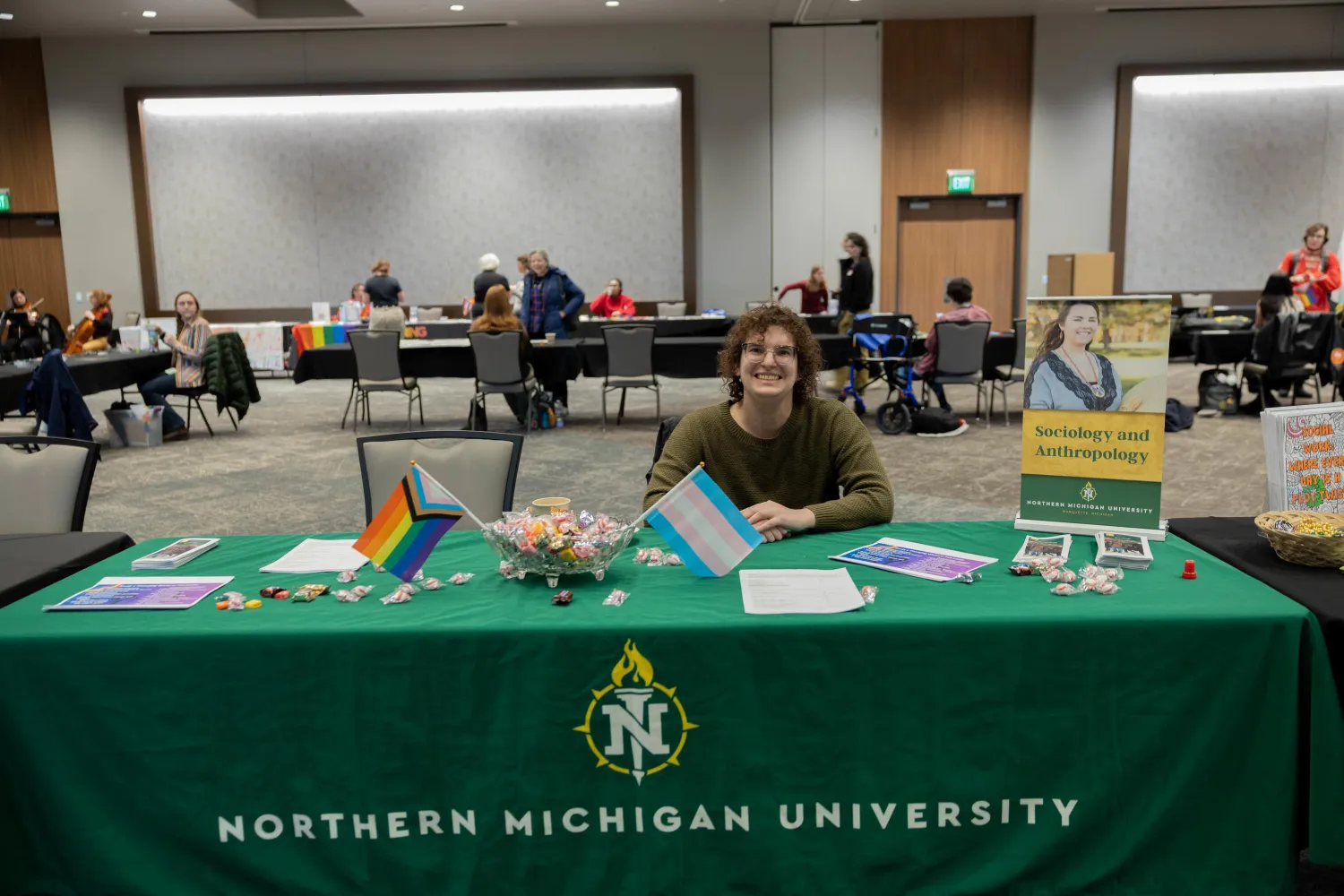 A person sits smiling behind a table with a green Northern Michigan University tablecloth, displaying LGBTQ+ flags, candy, and flyers at an indoor event with other tables and attendees in the background.