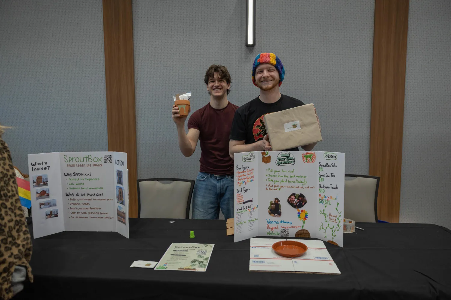 Two smiling people stand behind a table display about plant growth at an indoor event. One holds a potted plant, the other holds a brown paper bag. The table has posters, papers, and a small dish.
