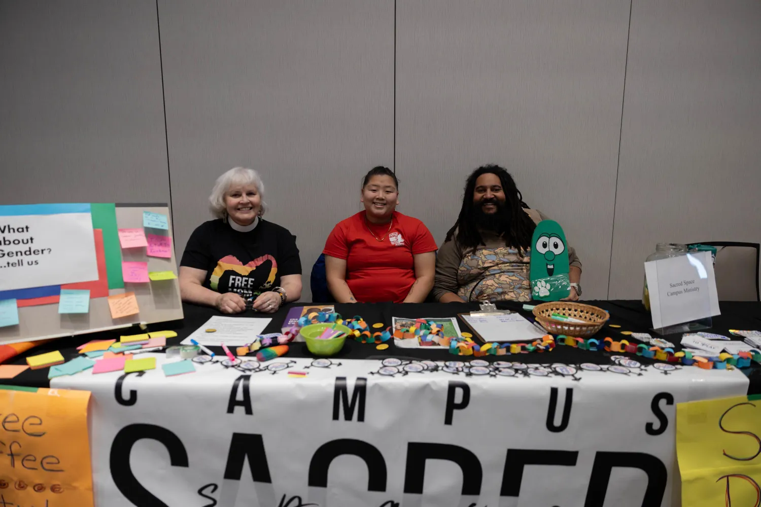 Three people sit behind a table covered with colorful items, forms, and signs at an event. A large "CAMPUS SACRED" banner hangs on the front, and a display board with sticky notes is on the left.