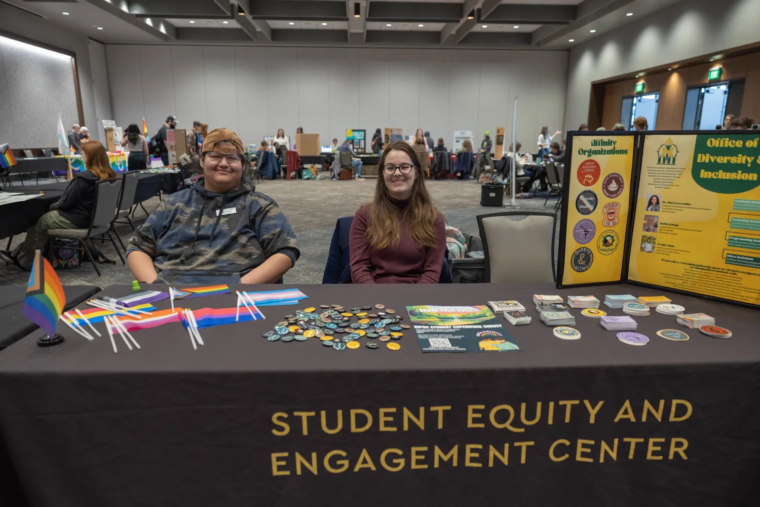 Two people sit behind a table labeled "Student Equity and Engagement Center," displaying LGBTQ+ flags, buttons, and informational materials at an indoor event. A diversity and inclusion board is visible beside them.