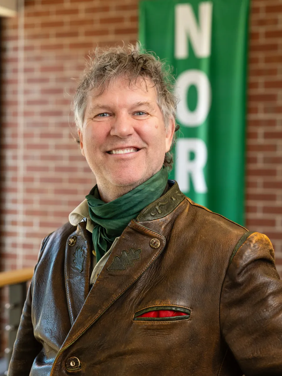 Man in leather coat and green scarf in front of brick wall