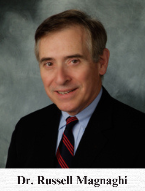 A younger Russell Magnaghi poses for a headshot against a grey background. He wears a crisp black blazer, baby blue undershirt, and a red and navy-striped tie.