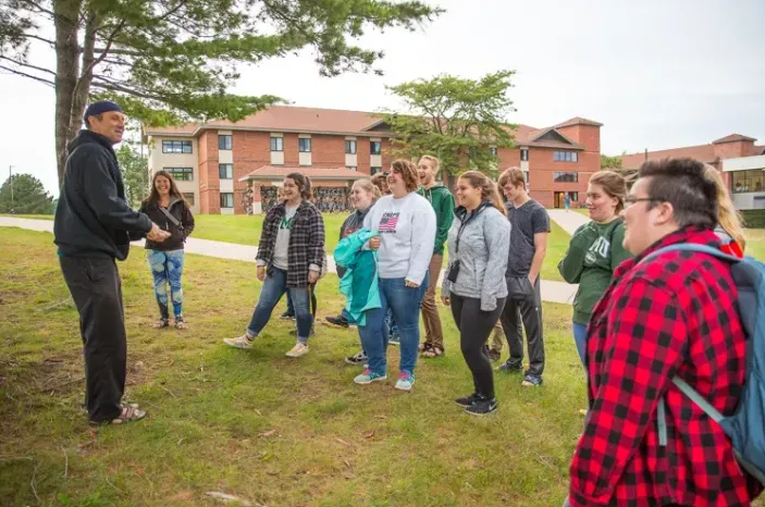 A group of NMU students stand on the lawn outside a dormitory and listen to an Anishinaabemowin lesson