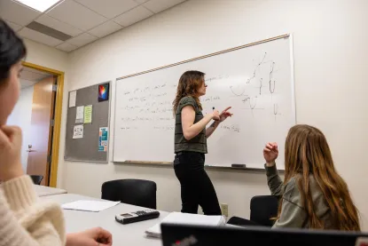 Student drawing mathematical graphs on a whiteboard while two students watch.