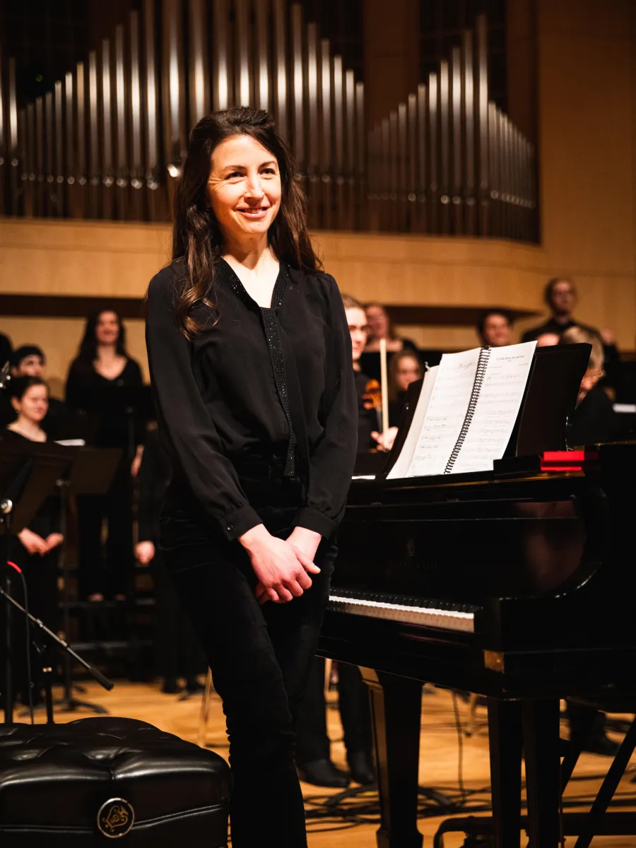 Dr. Theresa Camilli stands at a grand piano with members of a choir behind her.