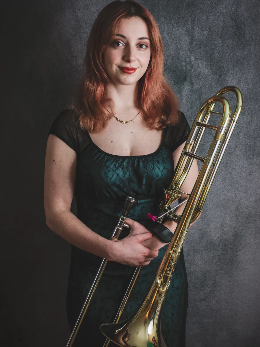 Arabella Olson stands against a dark gray studio backdrop, smiling while holding a trombone.
