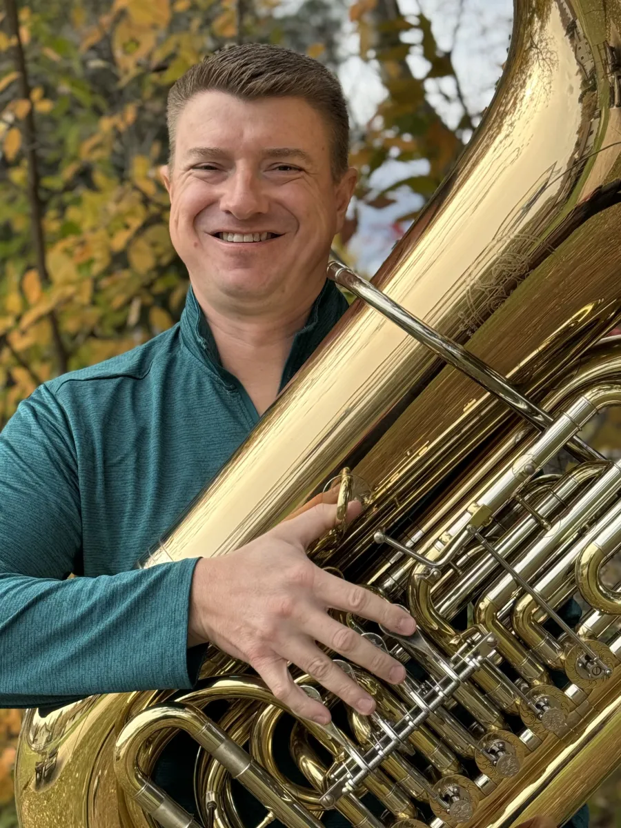 Smiling musician in a teal shirt holds a large polished brass tuba outdoors, with autumn trees behind him.