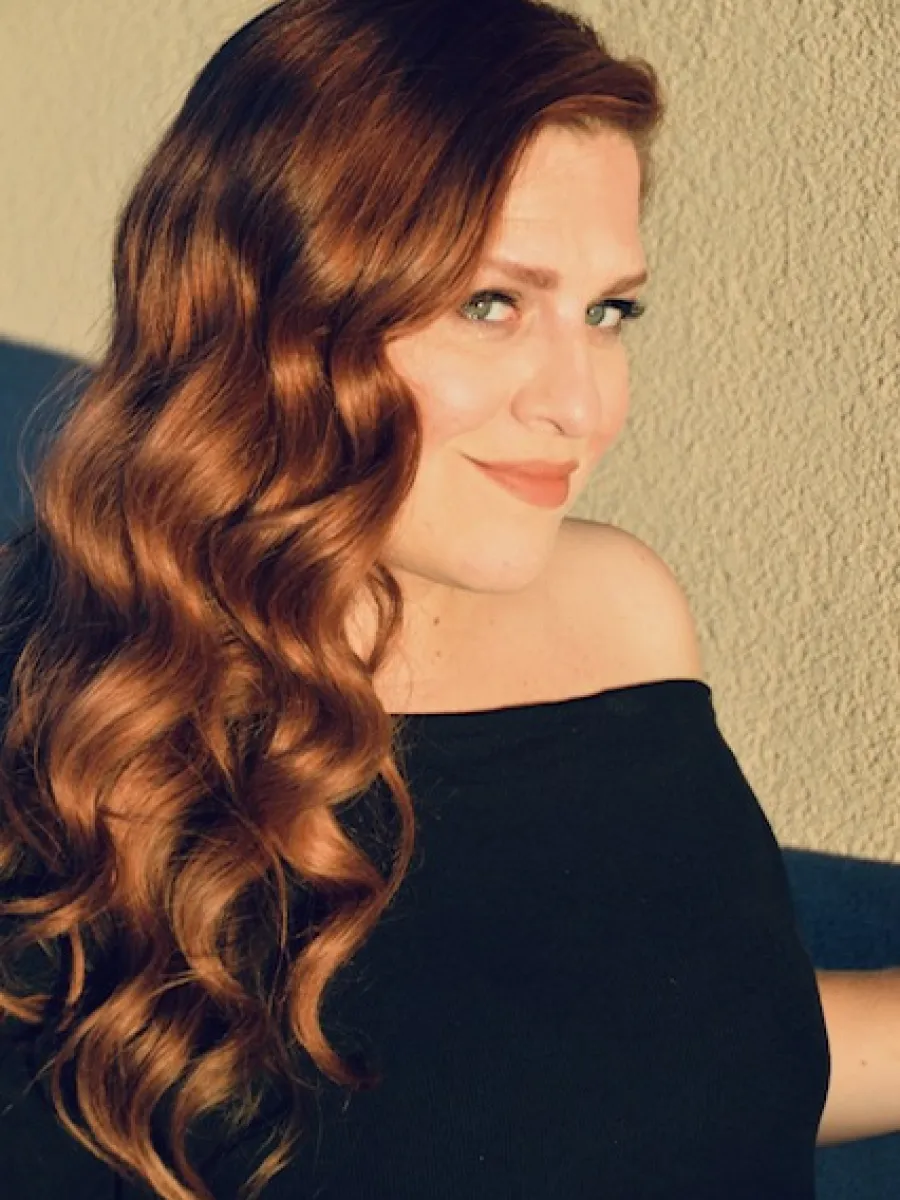 Portrait of a woman with long, wavy auburn hair in a black off-shoulder top, smiling at the camera in warm light.
