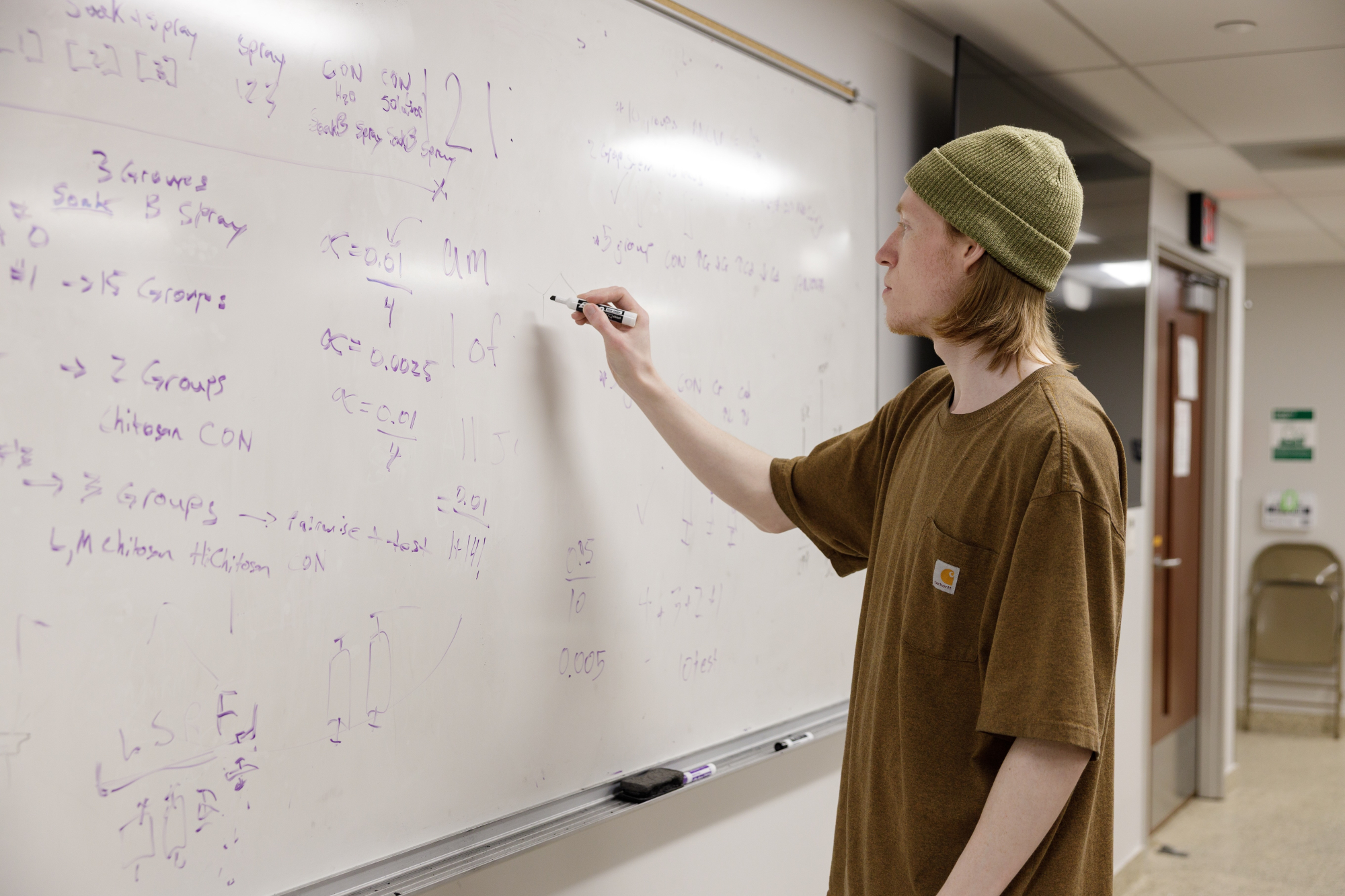 A student in a brown t-shirt and a green beanie writing on a whiteboard. The writing on the board is in purple marker with various math equations.