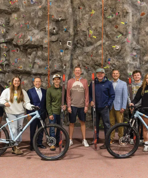Students and alumni supporters with bicycles, skis and backpacks with climbing wall in back
