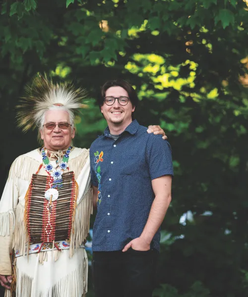 Two indigenous men, one in traditional dress, standing in front of a background of green leafed trees