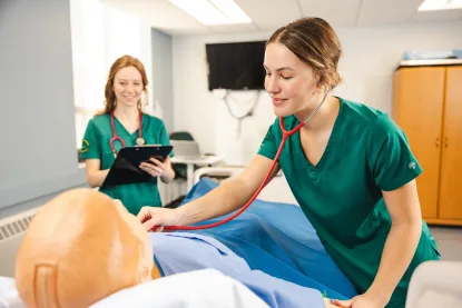 A nursing student checks the heartbeat of a mannequin in NMU's nursing simulation lab while another nursing student takes notes on a clip board