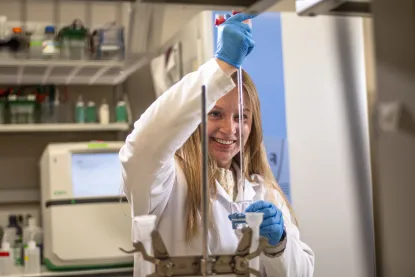 A student with long blonde hair, wearing blue gloves and a white lab coat pipes into a vial. The background is a lab setting with shelving of boxes and equipment on the counters lining the room.