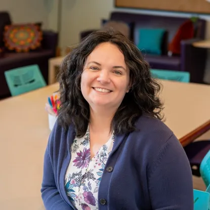 A portrait of an individual with dark, curly brown, shoulder length hair wearing a white and purple/pink floral shirt with a navy blue cardigan over top. They are sitting at a table in a classroom. Around the table, light teal chairs can be seen in the background. Subject is smiling at the camera.