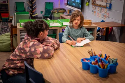 Two students sitting at a wooden table in a classroom with green bins behind them, a neon light in the corner and a projector screen on the wall. There is a box of coloring utensils in the middle of the table. One student is in a sage green shirt with a paper in their hands showing the other student something. The other student is in a side profile with their longer brown hair clipped with a white clip, wearing a brown fuzzy sweatshirt. 