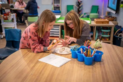 Two students sit at a wooden table with a stack of white paper and a bucket of coloring utensils in the middle. The students have a plate of objects between them. One student, with blonde hair and a pink sweater has a round, white ball in their hands an dis looking at the plate in front of them. The other student with blonde hair and glasses and a brown shirt is holding out their hands over the plate. 