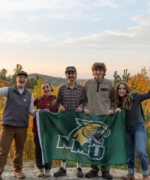 Five students holding a NMU flag, smiling at the camera, in front of fall foliage.