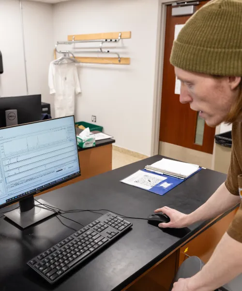 A student stands over a computer in a lab. Student is wearing a brown t-shirt with an olive green beanie