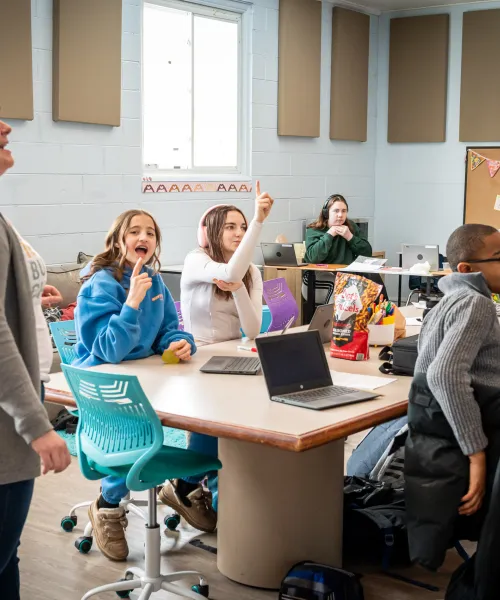 A classroom of students sitting at tables with laptops open. One table features a bag of pretzels. Students are engaged and smiling in the photo with a few looking directly at the camera and a few looking to the front of the classroom. Scattered students have headphones over their ears and one has their hand raised. The teacher is standing to the left in the photo slightly turned toward the students. They are in a white tshirt and a grey cardigan. They are taking with the students joyfully.