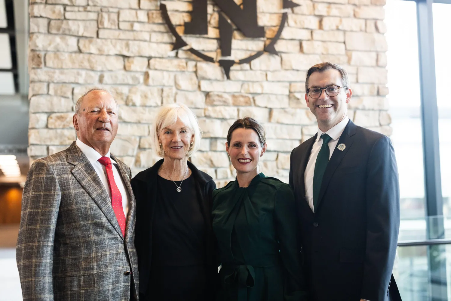 Four people stand together smiling in front of a stone wall with a large emblem. The group includes an older man in a plaid suit, an older woman in black, President Tessman's wife in green, and President Tessman in a suit and tie.