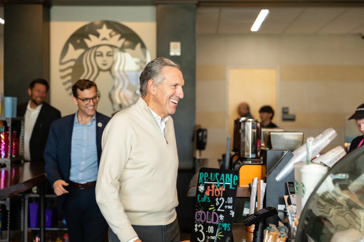 Howard Schultz, in a white sweater, smiles while standing at a counter in a cafe. President Tessman can be seen in the background, and there's a sign on the counter listing prices for hot and cold drinks. The Starbucks logo is visible on the wall behind him.