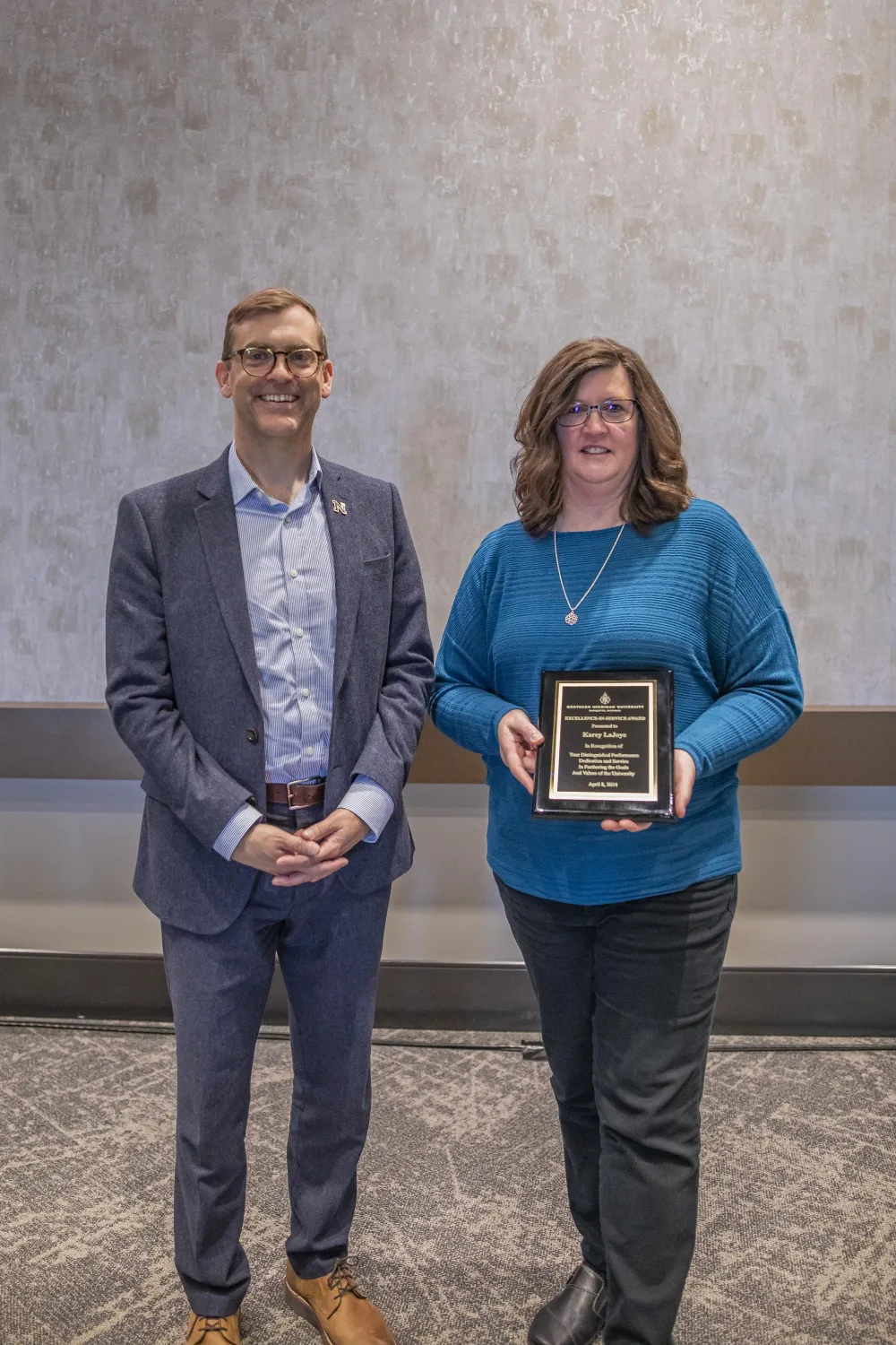 President Tessman stands next to a woman receiving an award. She is smiling and holding a plaque. Both are in front of a neutral background.