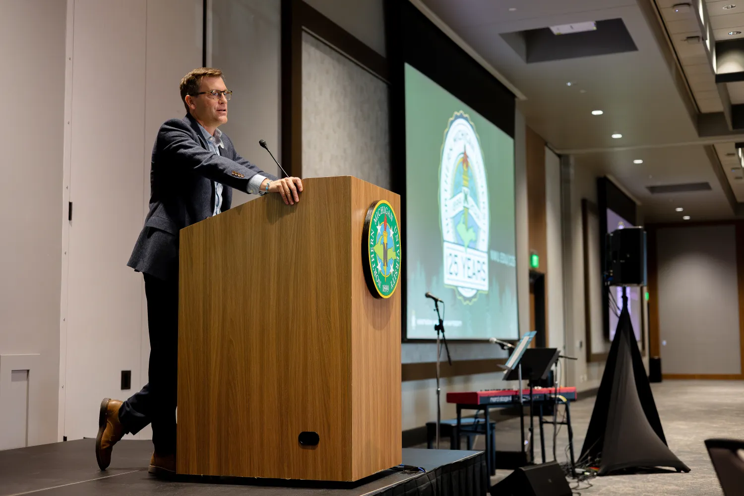 President Tessman, in a suit, stands at a wooden podium with a microphone, delivering a presentation. A large screen in the background displays the text "Convocation Address." Another sign is visible on the front of the podium.