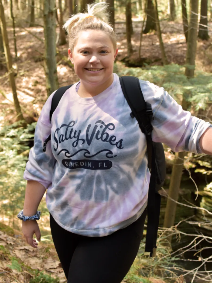 Woman smiling, hiking in a sunlit forest. She has a backpack and her hair in a bun, enjoying a peaceful moment.