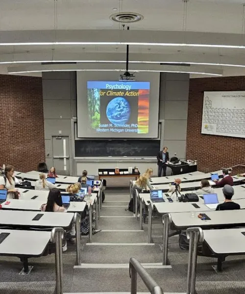 A lecture hall with tiered seating, partially filled with students using laptops. A large screen displays "Psychology for Climate Action."