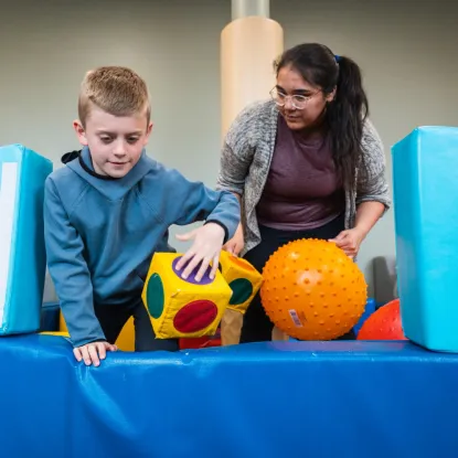 A young boy engages in play with colorful foam blocks and a spiky ball while a woman supervising smiles warmly, creating a vibrant, supportive environment.