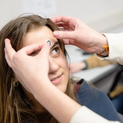 A person is gently placing a small, round sticker on a smiling woman's forehead. She appears curious and amused, with a background of a blurred room.