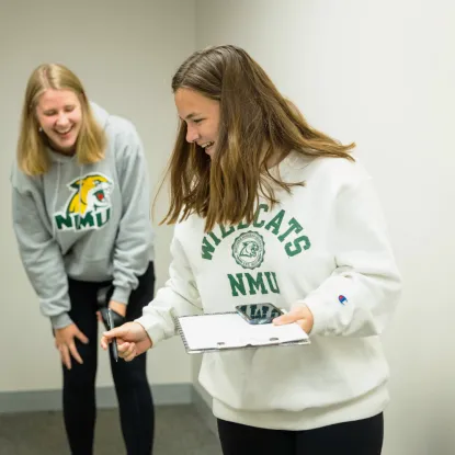 Two people in NMU Wildcat sweatshirts are laughing in a white room. One holds a clipboard and phone, conveying a playful, casual mood.