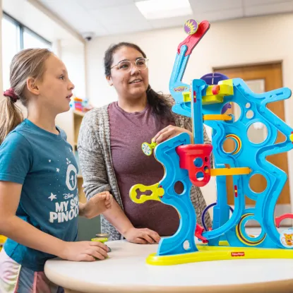A woman and a child stand at a table, engaged in play with a colorful blue and red marble run set. The scene is bright and cheerful.