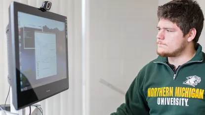 A person in a "Northern Michigan University" sweatshirt focuses intently on a computer screen displaying graphs and data, conveying concentration and analysis.