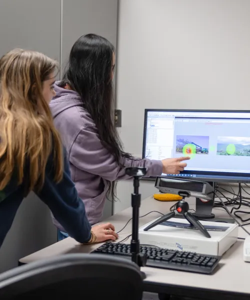 A group of people huddled around a computer in a research lab, engaged in a discussion. One person points at the screen showing colorful graphics.