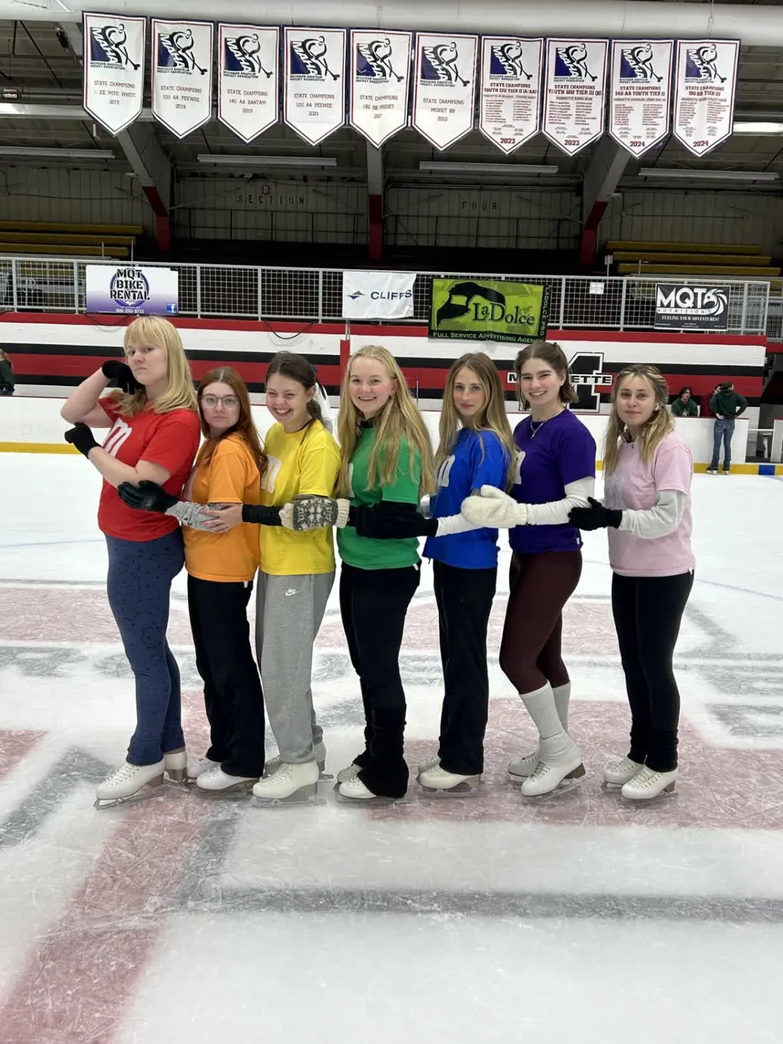 Seven skaters posing with shirts the color of the rainbow.