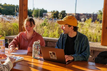Two students sitting at a picnic table in the sunshine, talking while working on laptops.