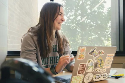 Woman with long brown hair wearing a Northern Michigan University hoodie on her laptop
