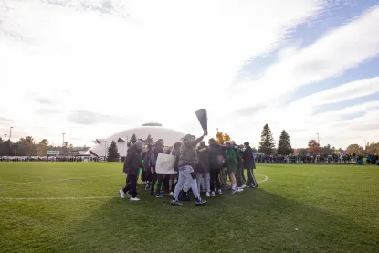 A group of people celebrate together on a grassy sports field under a bright sky, raising a large megaphone-like object, with trees and a domed building visible in the background.