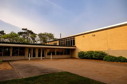 A modern, single-story building with large windows and tan brick walls, surrounded by bushes and trees, under a partly cloudy sky during sunset.