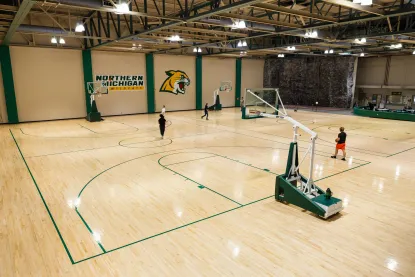 Wide view of an indoor basketball court with four hoops, labeled "Northern Michigan Wildcats." Three people are on the polished wood floor, and a rock climbing wall is visible in the background.
