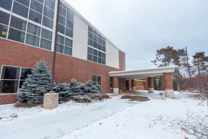 A brick and glass building with a covered entrance, surrounded by snow-covered ground and trees, on a cloudy winter day.