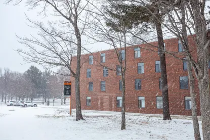 A red brick building with many windows stands amid leafless trees during a snowy winter day. Snow covers the ground, and parked cars are visible in the background near a black and orange campus sign.
