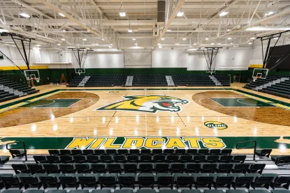 A brightly lit basketball court with "WILDCATS" and a wildcat logo at center court, green and yellow accents, and empty black bleachers on all sides. The gym has high ceilings and multiple basketball hoops.