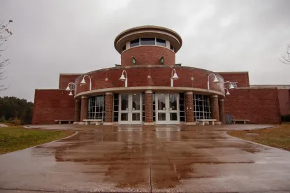 A round brick building with large windows, white pillars, and multiple white hanging lights at the entrance. The wet pavement reflects the structure on a cloudy, overcast day.