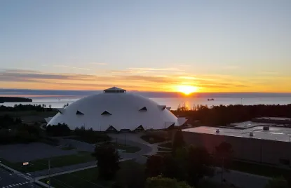 A domed stadium sits near a lake with a ship in the water as the sun rises, casting an orange glow over the horizon. Trees and buildings surround the stadium in the early morning light.